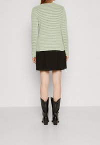 Světle zelený pruhovaný svetr s dlouhým rukávem v kombinaci s černou plisovanou sukní a černými kotníkovými botami, pohled zezadu.