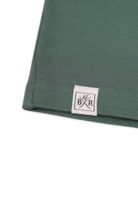 Grön bomullstyg med ribbad textur. En vit etikett med "B/R" och korsade redskap är sydd i fållen.