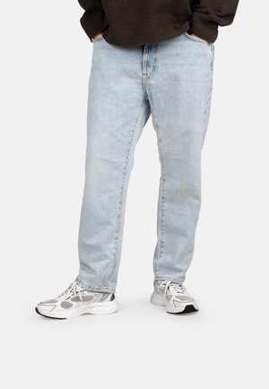 Person wearing light blue straight-leg jeans, white and gray sneakers, and a dark brown sweater with hands in pockets.