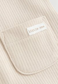 Un tissu côtelé beige présente une petite poche avec une étiquette blanche cousue, affichant le texte "little one" en marron clair.