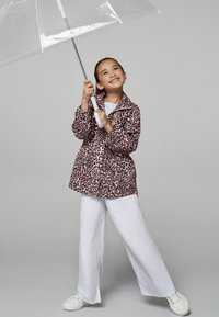 Regenjacke mit Leopardenmuster und großen Knöpfen, kombiniert mit weißen weit geschnittenen Hosen und weißen Turnschuhen, und hält einen durchsichtigen Regenschirm.