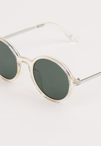 Pyöreät aurinkolasit, joissa on tummanvihreät linssit, kirkas muovikehys ja metalliset varret. Varusteena hienovarainen kaksoissilta.