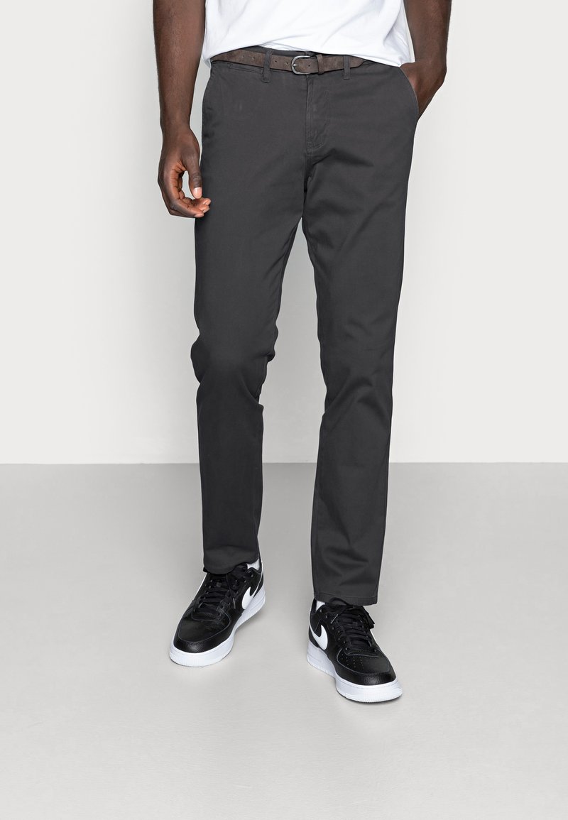 Homme portant un pantalon gris foncé, une ceinture noire, un T-shirt blanc et des baskets Nike noir et blanc, debout devant un fond clair uni.