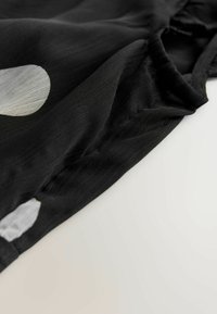 Schwarzer Stoff mit glatter Textur, der große, unregelmäßige weiße Punkte aufweist. Das Design umfasst einen gerafften Ausschnitt und Seitennähte.