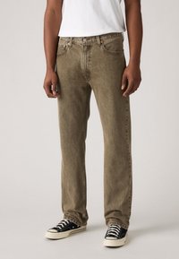 Jeans en denim vert olive avec une coupe droite, un design à cinq poches et une fermeture à un bouton, associés à des baskets noires et blanches.