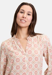 Blusa a maniche lunghe in beige chiaro con un motivo floreale rosa e blu. Presenta una chiusura con bottoni e spalle arricciate.