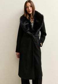 Cappotto nero con colletto in pelliccia morbida, dotato di vita rifinita da una cintura e design a fronte aperta. Il tessuto è liscio con una silhouette strutturata.