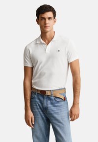 Man wearing white short-sleeve polo shirt with small chest logo, light blue jeans, and tan fabric belt with leather details.
