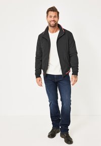 Schwarze Zip-Jacke mit roten und weißen Akzenten, kombiniert mit blauen Jeans. Strukturiertes Material, Stehkragen und taillierte Passform.