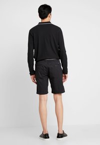 Camisa polo negra de manga larga combinada con pantalones cortos negros estampados. Los pantalones cortos tienen un diseño de puntos sutil y bolsillos laterales, usados con zapatillas negras.