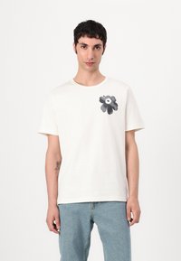 Vit bomull t-shirt med en mörk blommig grafik på vänster bröst. Korta ärmar och rundad halsringning. Lätt textur och avslappnad passform.
