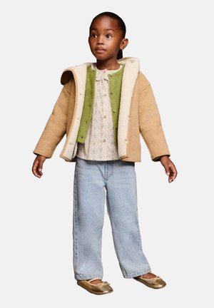 Chaqueta marrón claro forrada con borrego sobre un cárdigan verde acanalado; camiseta blanca con estampado, vaqueros de pierna ancha azul claro y zapatos planos dorados.