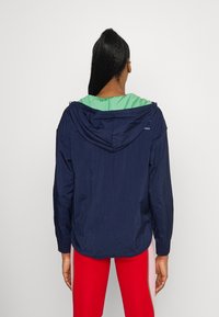 Lehký námořnický větrovka s zelenou kapucí, která zahrnuje stahovací kapuci a elastické manžety. Zadní pohled ukazuje hladkou strukturu.