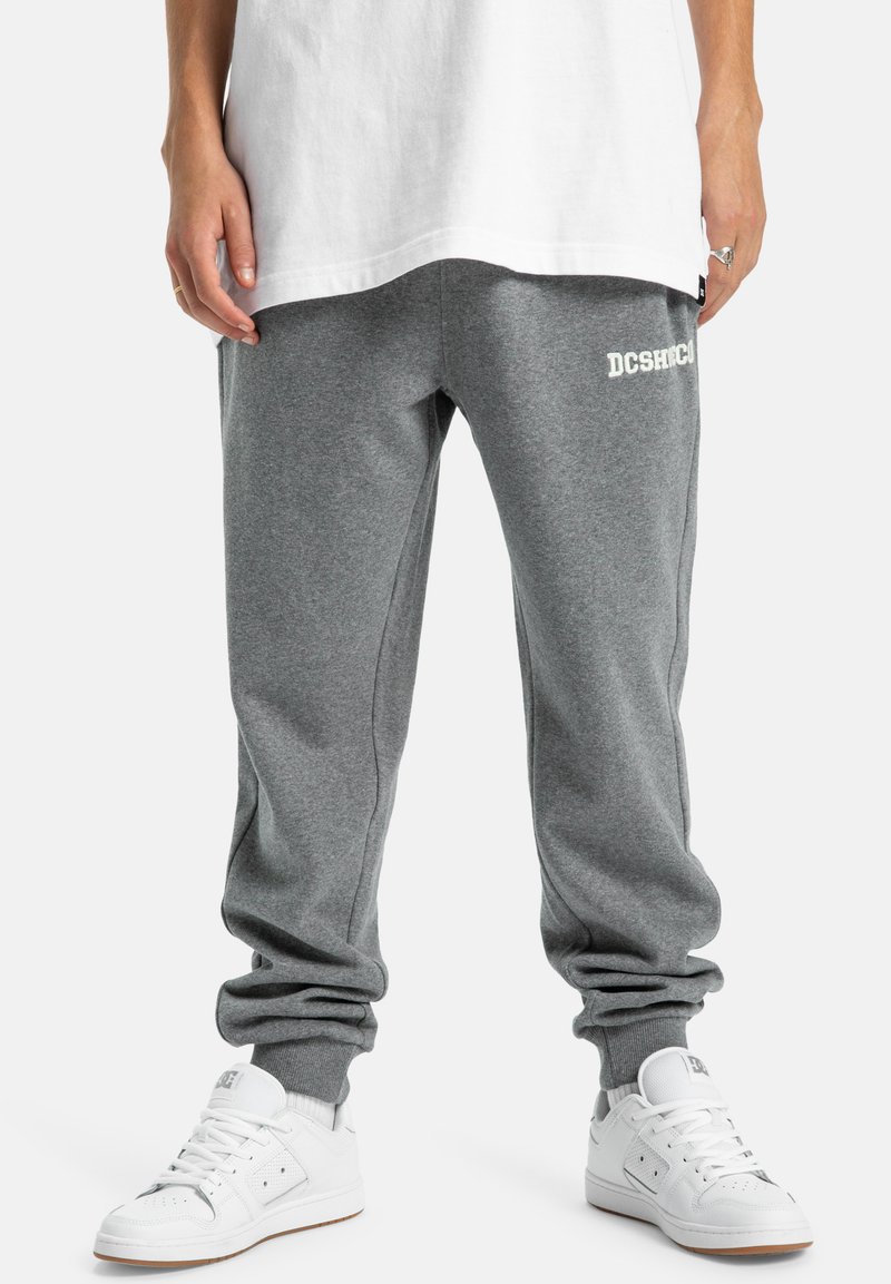 Graue Sweatpants mit Bündchen, die ein weißes Logo auf dem linken Oberschenkel haben. Kombiniert mit weißen Sneakers, die im Kontrast zur Textur des Stoffes stehen.