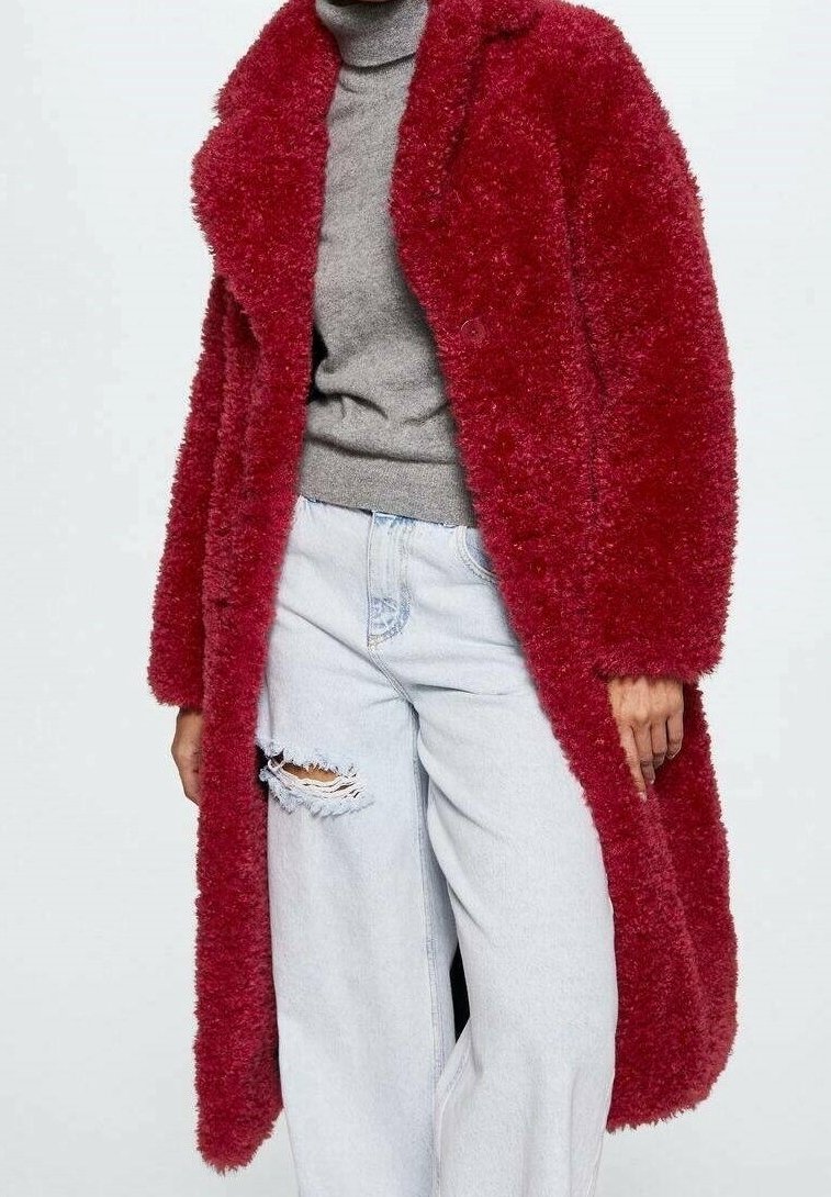 Personne portant un long manteau rouge texturé par-dessus un pull à col roulé gris et un jean bleu clair déchiré, sur un fond uni.