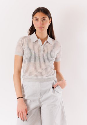 Donna con lunghi capelli scuri indossa una polo a maniche corte in maglia e pantaloni chiari a vita alta, in piedi con una mano in tasca.