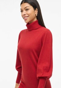 Vestido de punto rojo con un alto cuello de tortuga acanalado. Presenta mangas largas abullonadas con puños ajustados. Textura suave en toda la prenda.