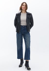 Černá kožená motorkářská bunda s ribovanou strukturou, béžový ribovaný top, modré džíny s vysokým pasem a třepeným lemem, a černé kotníkové boty na podpatku se zlatým řetízkem.