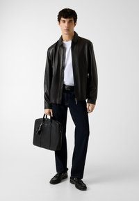 Zwarte leren jas, wit T-shirt, donkere spijkerbroek, houdt een zwarte tas vast. Heeft een gladde textuur; jas heeft een ritssluiting en kraag.