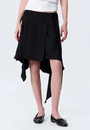 Femme portant une jupe portefeuille asymétrique noire à ourlet irrégulier, associée à des ballerines noires et un crop top blanc.