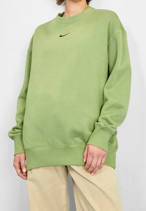 Osoba nosząca luźną, jasnzieloną bluzę Nike oraz beżowe spodnie, stojąca na jednolitym białym tle.