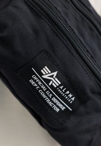 Musta nailonkangas, jossa on vetoketjullinen suljin ja etiketti, jossa lukee "Alpha Industries" sekä Yhdysvaltain puolustusministeriön urakoitsijateksti.