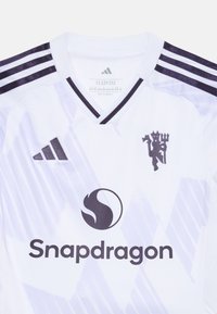 Hvit fotballtrøye med lilla detaljer, med en v-hals. Inkluderer mørk logo, teksturert mønster, og tre striper på skuldrene.