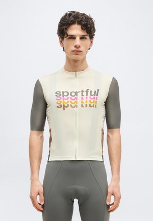 Jaunas vyras su garbanotais plaukais vilki smėlio spalvos ir pilką dviračių dresą su užrašu „sportful“ bei atitinkamus pilkus paminkštintus dviračių šortus.