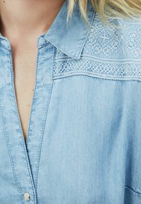 Light blue denim shirt with a collared neck, button placket, and white lace detailing across the shoulders. Soft texture, casual design.
