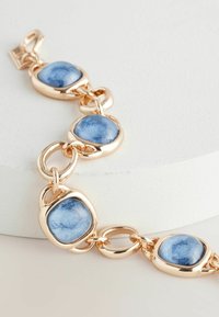 Bracelet en tone or avec des pierres en émail bleu ovales, interconnectées par des maillons lisses et brillants. Pierres texturées dans des montures en métal poli.