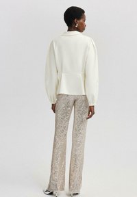 Biela štruktúrovaná bunda s nafúknutými dlhými rukávmi a peplum detailom, kombinovaná s lesklými béžovými trblietavými nohavicami, vytvára elegantný siluetu.