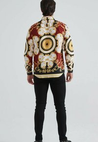 Un homme debout de dos, portant une chemise à manches longues avec des motifs baroques ornés rouges, dorés, noirs et blancs, ainsi que des pantalons noirs ajustés.