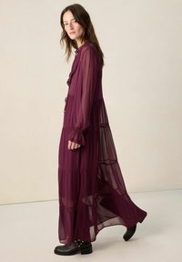 Robe longue et transparente couleur bordeaux avec un col volanté et un design à étages ; munie de manches longues et de poignets élastiques ; portée avec des bottines noires épaisses.