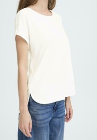 Kvinde iført en løs hvid t-shirt med korte ærmer og blå jeans, stående mod en ensfarvet lys baggrund.