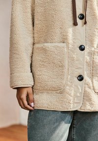 Giacca di pile beige con una texture morbida, dotata di bottoni neri, un laccio marrone e due tasche frontali.