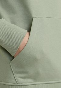 Sweatshirt vert clair avec des poignets côtelés et une poche avant, à la texture lisse et aux coutures nettes. Design simple et décontracté.