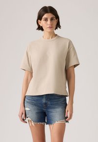 Γυναίκα με κοντά σκούρα μαλλιά που φοράει μπεζ φαρδιά μπλούζα και σκισμένο μπλε τζιν σορτς, μπροστά σε απλό φόντο.