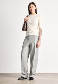 Jasnokremowy sweter z krótkim rękawem, szare plisowane spodnie i czarne buty z noskiem w szpic. Modelka niesie ciemnobrązową skórzaną torbę.