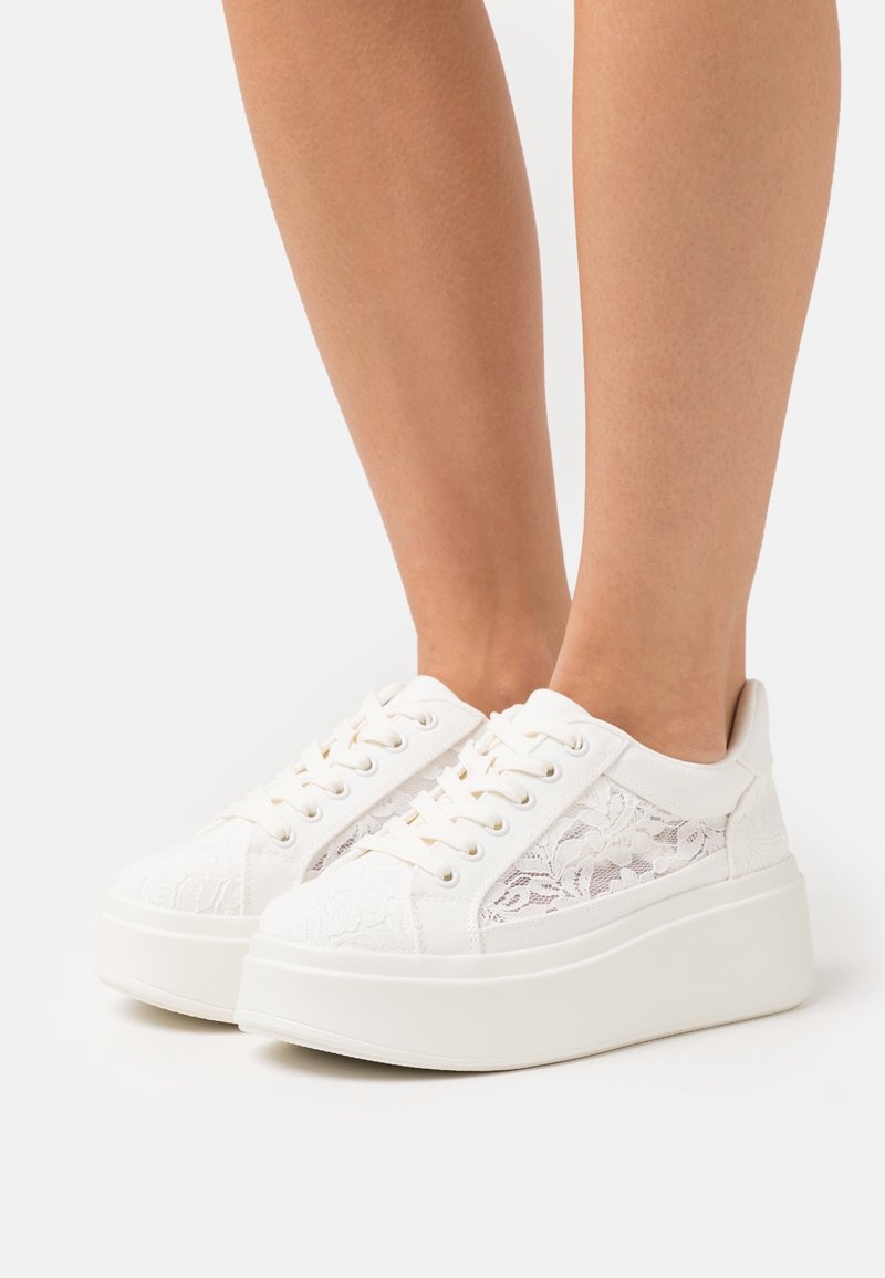Baskets à plateforme blanches avec motif floral en dentelle portées pieds nus sur un fond blanc uni.