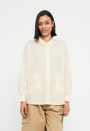 Chemise beige avec un col à revers, une fermeture boutonnée à l'avant et de longues manches bouffantes. Comprend deux poches décoratives aux motifs géométriques.
