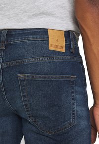 Vaqueros azul oscuro con un acabado de mezclilla texturizada. Presenta un parche de cuero visible etiquetado "ONLY & SONS" en la cinturilla.