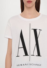 White cotton t-shirt featuring a bold black "AX" logo design with vertical lines and the text "ARMANIEXCHANGE" below. Short sleeves.