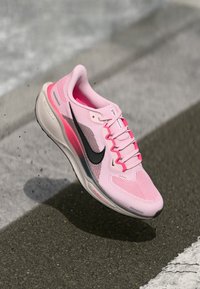 Svetloružová športová obuv s priedušným sieťovaným zvrškom, čiernym Nike swoosh, ružovými akcentmi a tlmenou podrážkou, ktorá sa vznáša nad sivým povrchom.