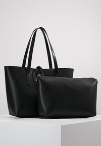 Sac fourre-tout en cuir noir avec de longues poignées, accompagné d'une pochette assortie à fermeture éclair, tous deux présentés sur un piédestal blanc devant un fond gris.
