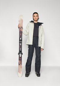 Skidutrustning med en ljusbeige jacka, svarta byxor, en mörk tröja och ett par skidor med skidhållare, placerad mot en neutral bakgrund.