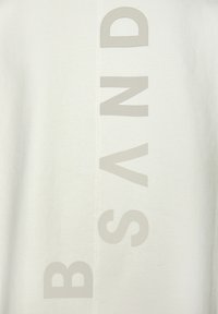 Weiße Baumwoll-T-Shirt mit großen hellgrauen Buchstaben, die "B S A N D" vertikal in der Mitte aufdrucken, mit sichtbaren Nähdetails.