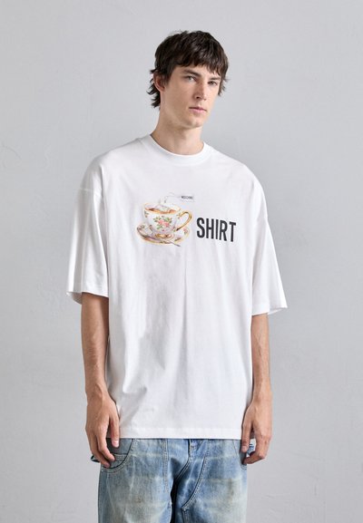 Witte oversized t-shirt met een gedrukte afbeelding van een theekopje en schotel, met het woord "SHIRT" in vetgedrukte zwarte tekst. Katoenen materiaal, ontspannen pasvorm.