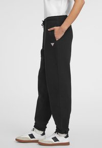 Zwarte trainingsbroeken met elastische manchetten, een trekkoord in de taille, zijzakken en een klein driehoekig logo op de linker bovenbeen. Gepaard met sneakers.