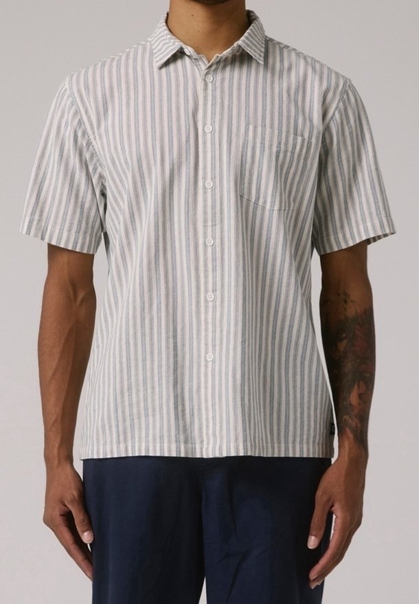 Homme portant une chemise à manches courtes boutonnée avec des rayures verticales grises et blanches et un pantalon bleu marine.
