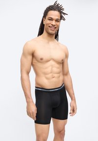 Άνδρας χωρίς πουκάμισο με μακριά dreadlocks, όρθιος και χαμογελαστός, φοράει μαύρα μπόξερ Calvin Klein σε απλό φωτεινό φόντο.